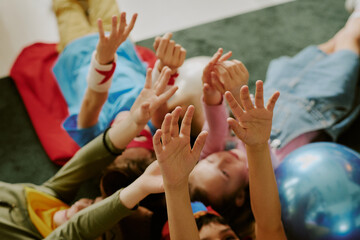 Group of children lying on floor reaching hands upward, diverse boys and girls engaging in playful activity together, visible faces and arms showing excitement and interaction