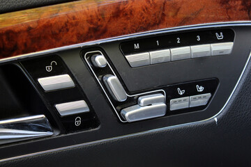Close-up of luxury car door seat adjustment controls in silver and black. Buttons include seat position, ventilation, and heating options. Set in a black plastic and leather panel with wood trim
