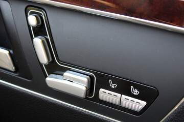 Close-up of luxury car door seat adjustment controls in silver and black. Buttons include seat position, ventilation, and heating options. Set in a black plastic and leather panel with wood trim