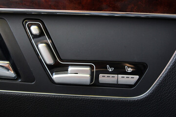 Close-up of luxury car door seat adjustment controls in silver and black. Buttons include seat position, ventilation, and heating options. Set in a black plastic and leather panel with wood trim