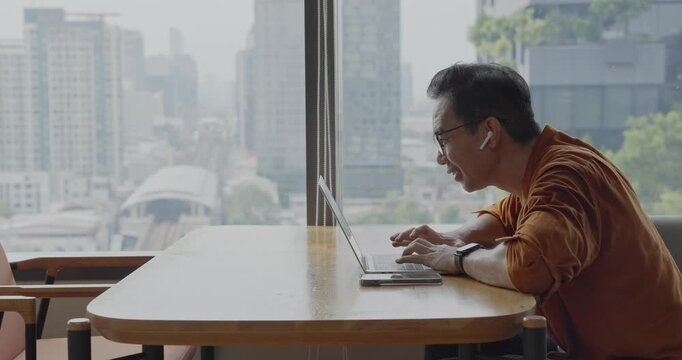 Senior Asian businessman wearing headset celebrates success during online meeting in modern co-working space. Remote work and business communication concept	
