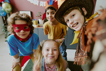 Group of multiethnic children smiling and wearing costumes at party, looking toward camera, including Caucasian boy in cowboy hat, biracial boy in feathered headband, girl with party hat