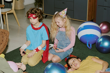 Group of children sitting and lying on floor during birthday party, Caucasian boy and girl wearing party hats and costumes, biracial boy lying nearby, all interacting together