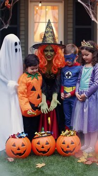 A green-faced witch with vibrant orange hair and a black hat gives candy to four diverse children in ghost, pumpkin, superhero, princess costumes on a decorated porch with jack-o'-lanterns and mist.