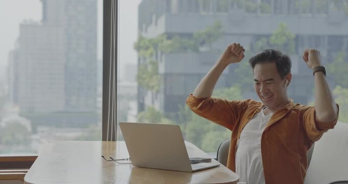 Senior Asian businessman wearing headset celebrates success during online meeting in modern co-working space. Remote work and business communication concept	
