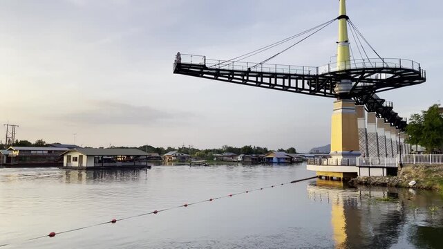 Riverside skywalk in Kanchanaburi, Thailand, extending over the water with scenic views of floating houses and riverboats in the background