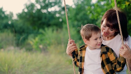 Mother, little son swing on swing in park under tree at sunset. Happy family, kid boy, swing. Concept of family happiness, dreams, entertainment. Child, mother plays on wooden swing, dreams of flying