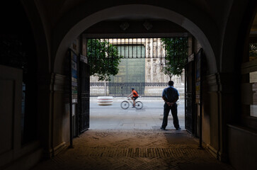A man stands under an archway, looking out at a street with a blurred cyclist passing by, framed by intricate architecture in the distance.