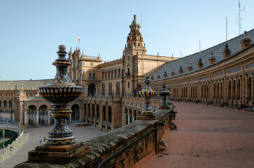 A high-angle shot captures the stunning architecture of the Plaza de España in Seville, Spain. The focus is on the ornate ceramic balustrade in the foreground, leading the eye towards the building