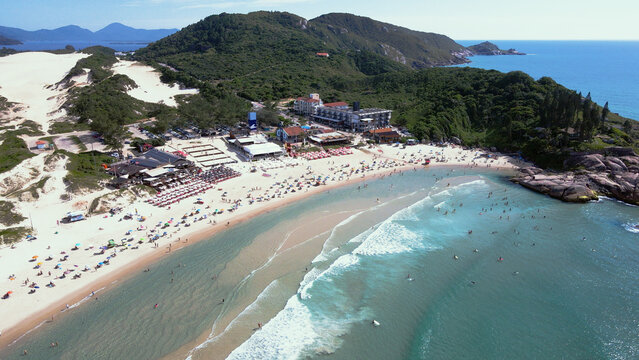 Panoramic aerial view of Joaquina Beach in Florianopolis, Santa Catarina, crowded with people and umbrellas on a sunny summer day with turquoise blue sea.
