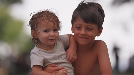 Smiling siblings embracing outdoors, older brother holding younger baby sister with affectionate arm around her, cheerful childhood bonding and happy family summer moment