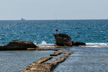 A person fishing while sitting on a rock formation in the Mediterranean Sea in Acre, Akko, Israel on a sunny day.
