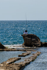 A person fishing while sitting on a rock formation in the Mediterranean Sea in Acre, Akko, Israel on a sunny day.
