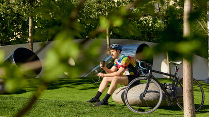 Cyclist in colorful jersey drinking coffee during rest on green lawn with relaxed mood. Concept of...