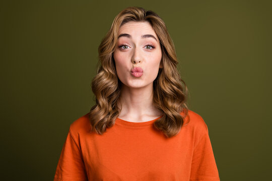 Charming young woman with curly blonde hair in an orange t-shirt smiling playfully on a green background - Powered by Adobe