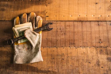 Fotobehang Brandhout Leather work gloves holding a rusty wrench and American dollars on a rustic wooden work table  © roy
