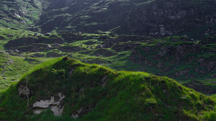 Green grass grows on rolling hills in the Gap of Dunloe, County Kerry, Ireland. Rocks are found on hillsides. The sun shines on the landscape.