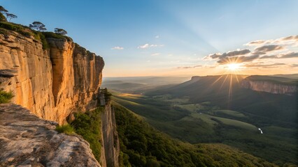 The golden sunset casts long rays over a magnificent valley, showcasing towering bluffs and lush green forests below a clear blue sky.