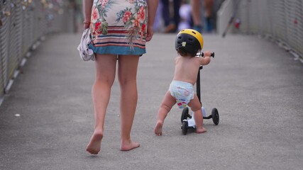 Toddler riding scooter with yellow helmet on bridge while mother walks barefoot nearby, capturing independence, learning, and early childhood exploration in outdoor setting
