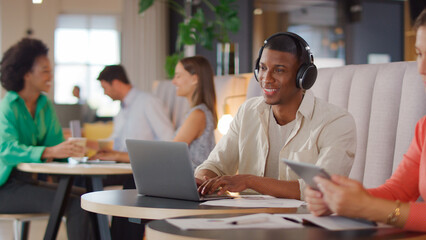 Businessman Wearing Wireless Headphones Working On Laptop In Informal Seating Area Of Modern Office