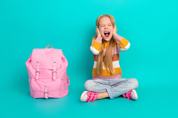 Laughing young girl sitting cross-legged on a turquoise background with a pink backpack, expressing a playful cheerful mood