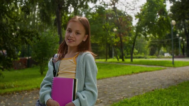 Portrait shot of young girl carrying backpack and pink notebook, standing on paved path in sunny park