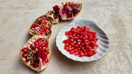 Loose pomegranate (Punica granatum) seeds in a white ceramic plate shot on a wood table. There are pieces of ripe pomegranate fruit around it. Pomegranates are a super food.