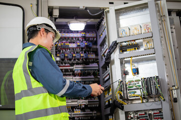 Worker wearing safety helmet and vest performs inspection at high voltage substation control panel...