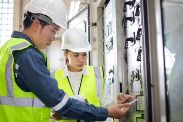 Worker in high visibility vest and helmet performing substation control inspection with another worker, focusing on high voltage equipment and safety procedures in industrial environment