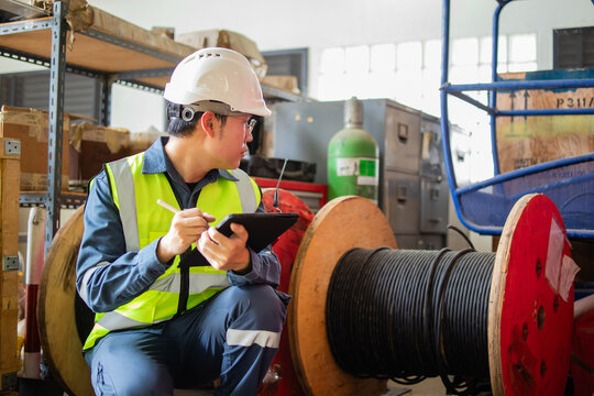 Worker wearing safety helmet and vest conducts inspection of power cable in industrial warehouse environment using digital tablet for documentation and quality control