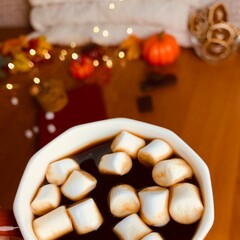 Sipping a warm drink topped with toasted marshmallows in a cozy setting filled with autumn decorations. Soft lights and seasonal elements enhance the atmosphere