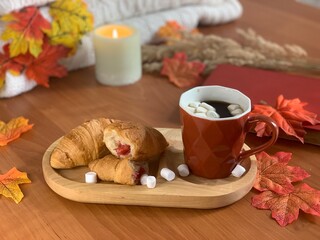 Enjoy a warm autumn atmosphere with a wooden tray displaying croissants filled with fruit, a steaming mug topped with marshmallows, and colorful leaves surrounding the setting