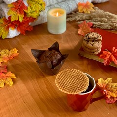 Enjoy delightful autumn flavors with a chocolate muffin, a cookie, and a waffle atop a cozy table adorned with colorful leaves and a candle, creating a warm atmosphere