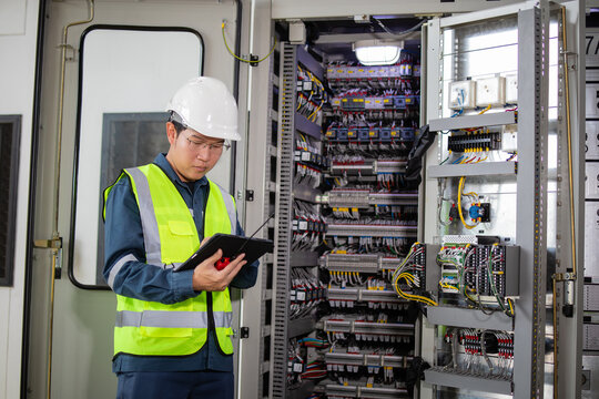 Inspector wearing safety helmet and high visibility vest is controlling high voltage energy system using tablet in industrial control room with complex wiring and equipment - Powered by Adobe
