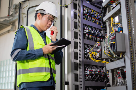 Inspector in high visibility vest and helmet controls energy system using tablet and radio near complex high voltage electrical control panel with numerous wires and components