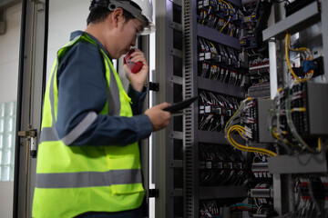 Electrical engineer wearing safety helmet and reflective vest performing inspection at substation control panel using tablet and walkie talkie for communication and safety check