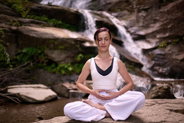 Young woman sitting in lotus pose under tropical waterfall meditating with calm strength and mindfulness fully connected to nature energy tranquility and inner balance