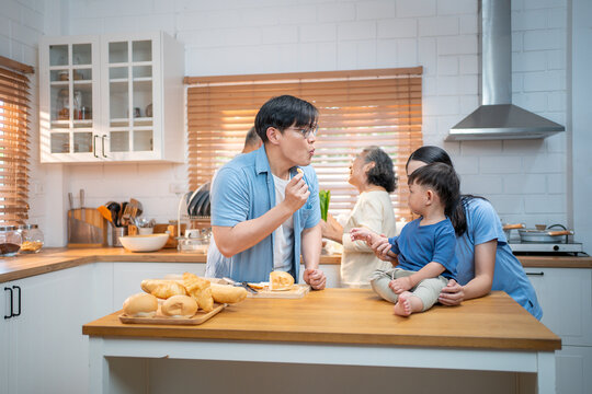 Asian family preparing food together in a cozy kitchen  a father slicing bread while the mother and child watch joyfully, surrounded by three generations. - Powered by Adobe