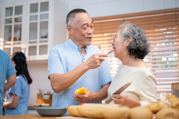 Happy Asian senior couple preparing fruit together in the kitchen while sharing laughter and love, surrounded by family in the background. A heartwarming moment of connection and healthy living.