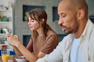 Young woman browsing her phone while enjoying breakfast with her husband at home, creating tension as he looks visibly annoyed