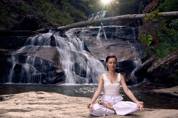 Young woman sitting in lotus pose under tropical waterfall meditating with calm strength and mindfulness fully connected to nature energy tranquility and inner balance
