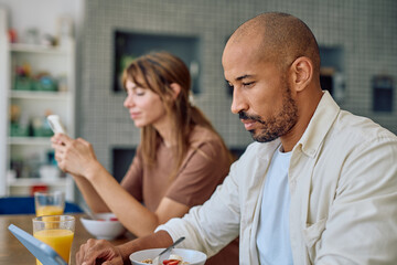 Interracial couple ignoring each other while using their electronic devices during breakfast in the kitchen
