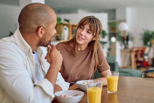 Multi-ethnic couple having breakfast at home, eating cereals and drinking orange juice while talking and smiling