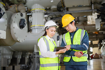 Two engineers in safety gear collaborate at electrical substation, discussing plans on tablet....