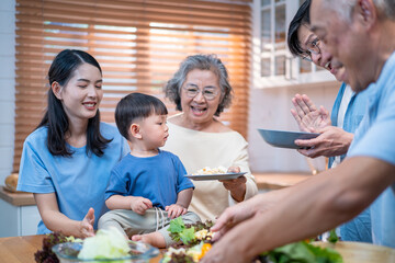 Naklejka premium : Happy multi-generational Asian family preparing salad together in the kitchen. A young child participates by holding a tomato, highlighting warmth, healthy lifestyle, and family bonding.