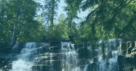 Cascading waterfall flowing over rock ledges in mountain forest, with moss patches and evergreens