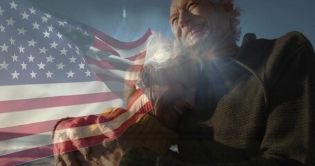 Fototapeta premium Smiling senior couple embracing on sandy beach, wearing warm jackets with American flag overlay