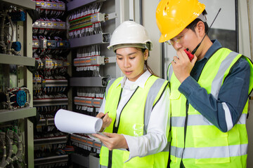 Two engineers in safety gear collaborate in industrial setting, examining documents and discussing electrical systems. They are focused and engaged in their work