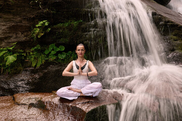 Young woman sitting in lotus pose under tropical waterfall meditating with calm strength and mindfulness fully connected to nature energy tranquility and inner balance