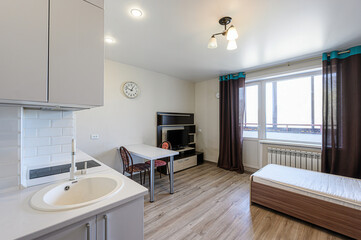 bright studio apartment features a kitchen area with white cabinets, a dining table, a TV stand, and a bed by the window. Brown curtains with teal accents add a touch of color